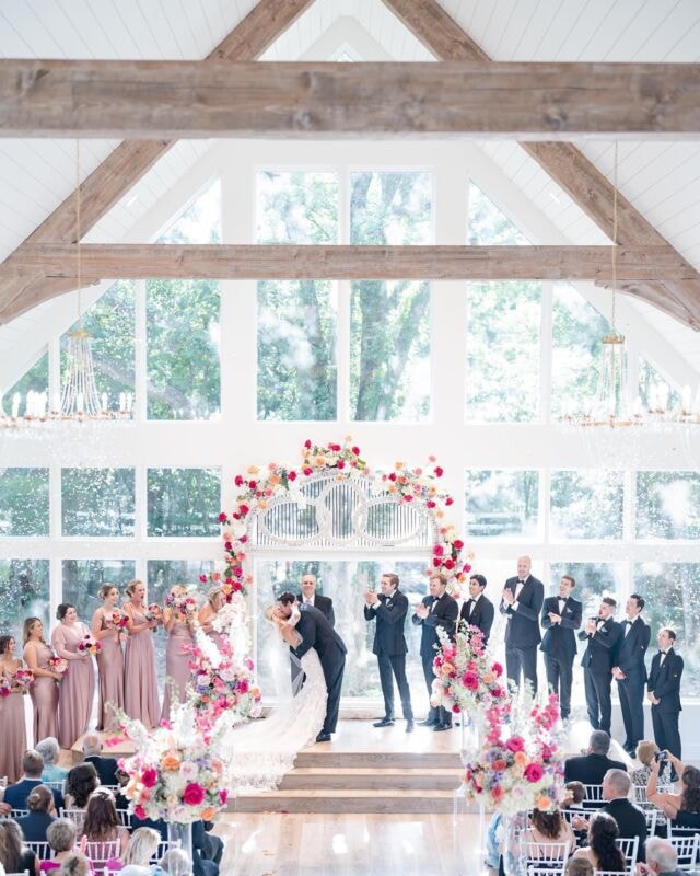 Couple sharing their first kiss at the altar inside the French Farmhouse chapel with a vibrant colorful floral arch and cheering wedding party