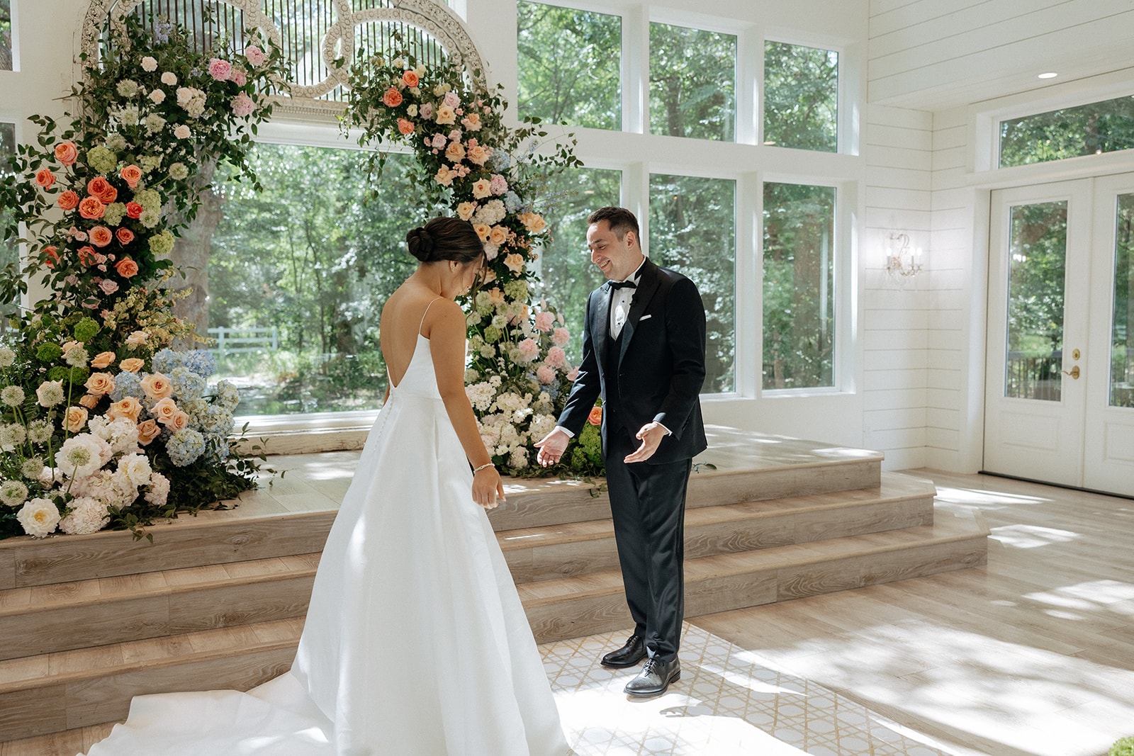 Drew's first look reaction as Ally walks toward him in her strapless A-line gown with a cathedral train, in front of the chapel's towering floral arch of coral, peach, blue, and white blooms with the floor-to-ceiling windows behind them