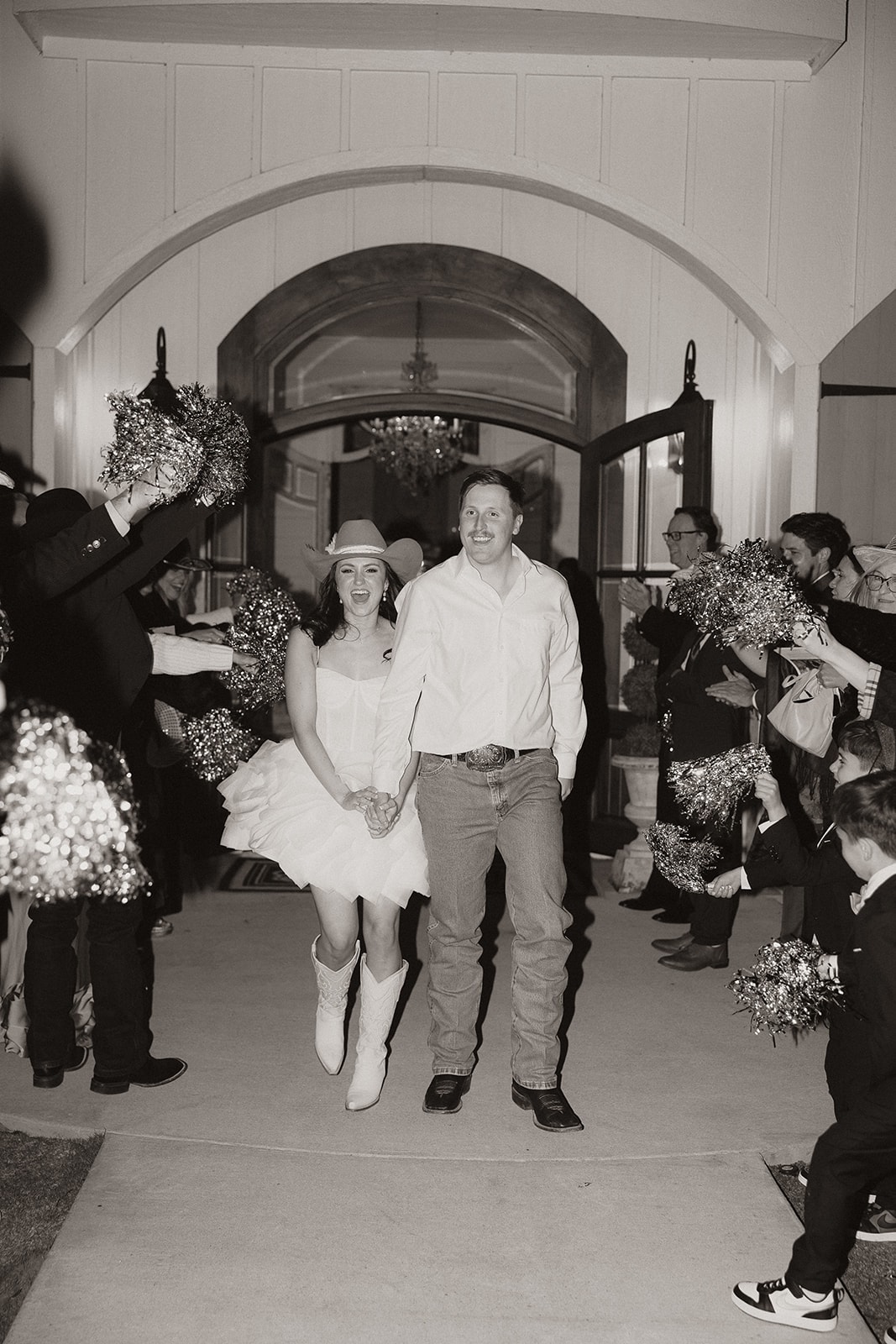 Black and white photo of Avery and Pearson running through a pom-pom send-off at the venue entrance