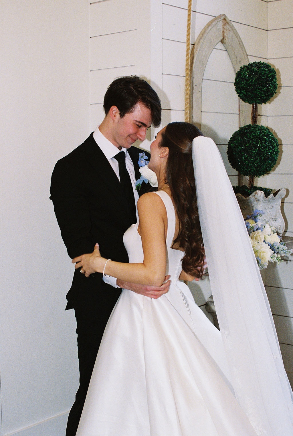 Brooke beaming and holding her blue delphinium and white bouquet high as Shepard kisses her cheek during their recessional down the chapel aisle, guests cheering on both sides