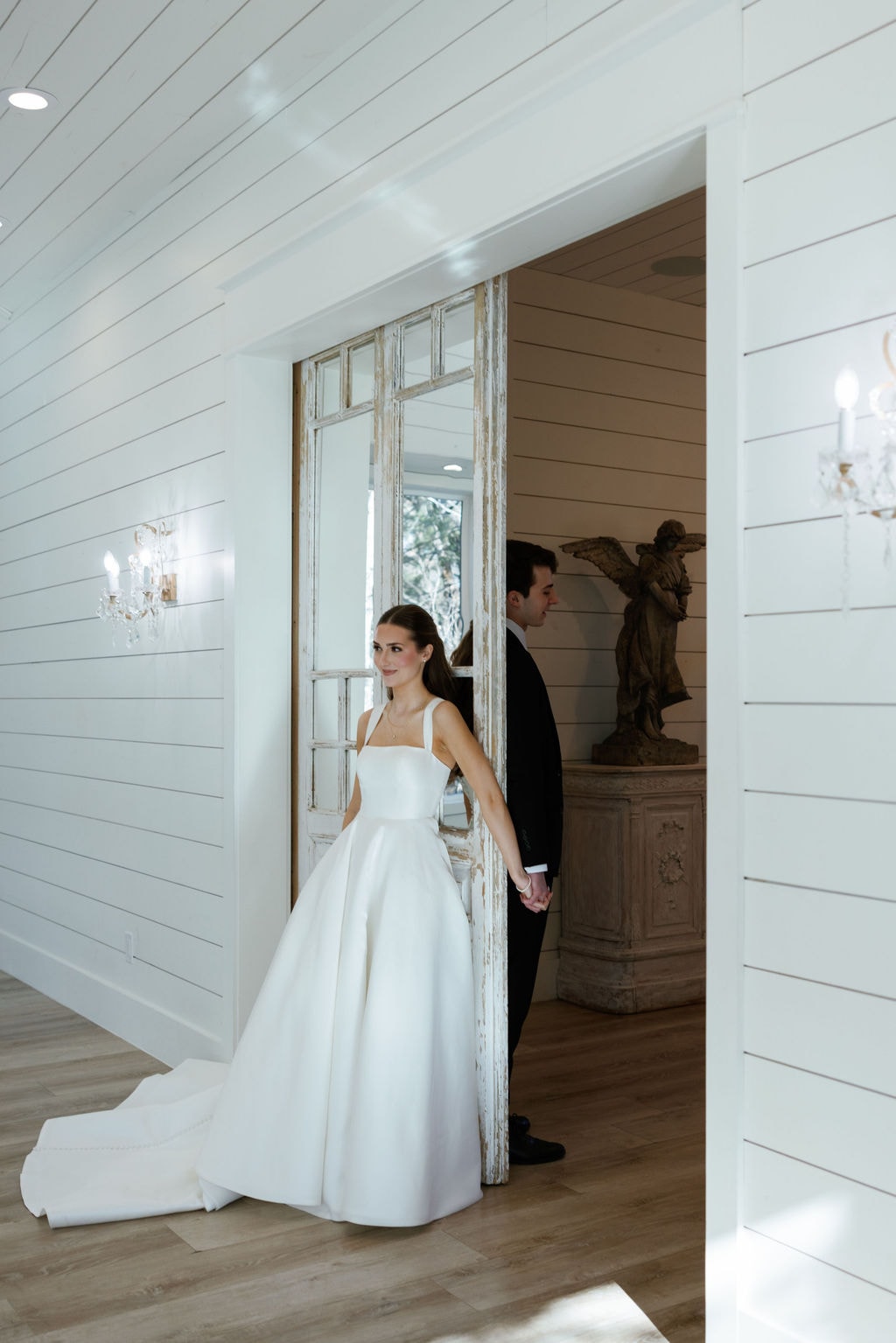Wide shot of Brooke and Shepard holding hands through the antique French doors — Brooke smiling softly in her white ballgown, Shepard in a black tuxedo facing away, the angel statue and crystal sconces visible in the background
