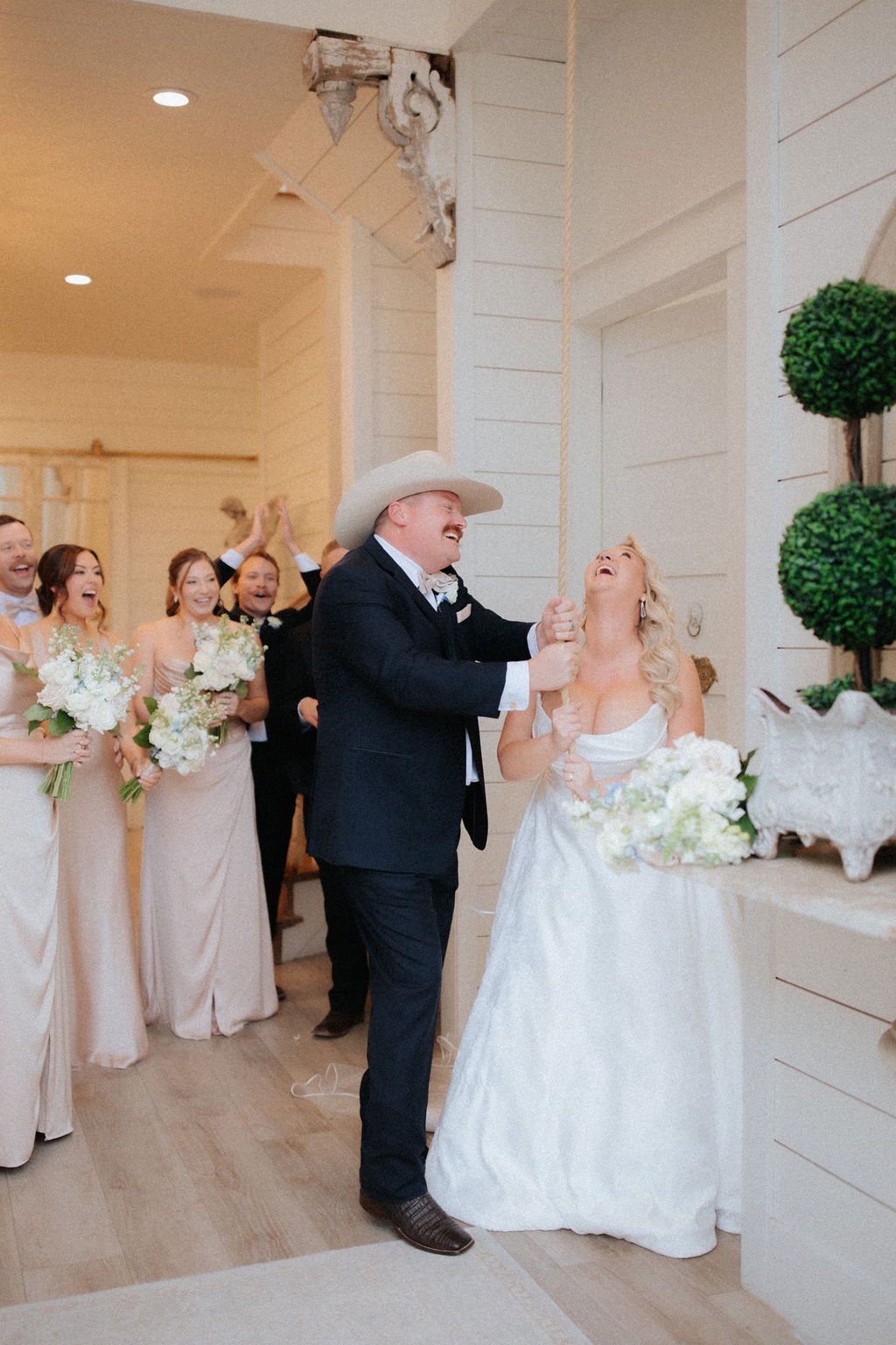 Kara and Anthony Marquardt laughing together as they ring the chapel bell after their ceremony, surrounded by their cheering bridal party at The French Farmhouse Venue