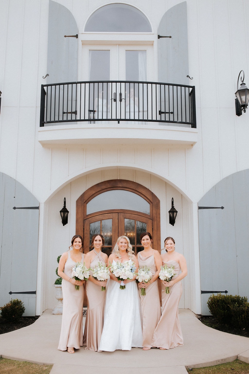 Kara and her four bridesmaids in blush satin gowns holding white and blue bouquets, standing in front of the grand arched entrance doors of The French Farmhouse Venue
