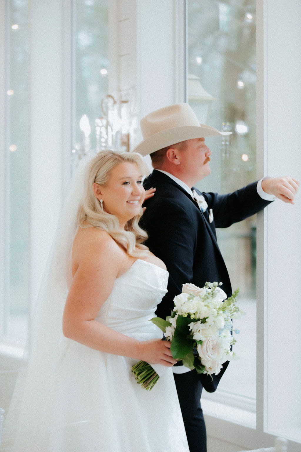 Kara and Anthony Marquardt sharing their first look — Kara in her strapless white ballgown holding a white and blush bouquet, Anthony in a navy suit and cream cowboy hat, standing together by the floor-to-ceiling windows of The French Farmhouse reception hall