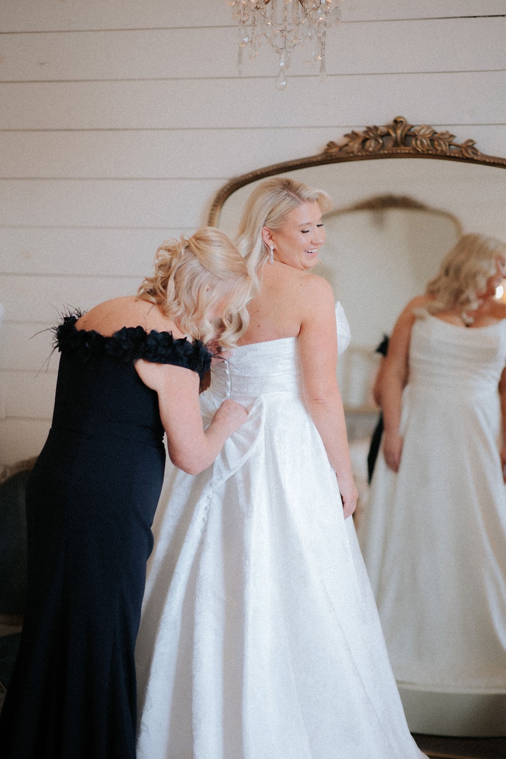 Kara smiling as her mom buttons the back of her strapless white ballgown in the bridal suite at The French Farmhouse Venue, reflected in the ornate gilded mirror