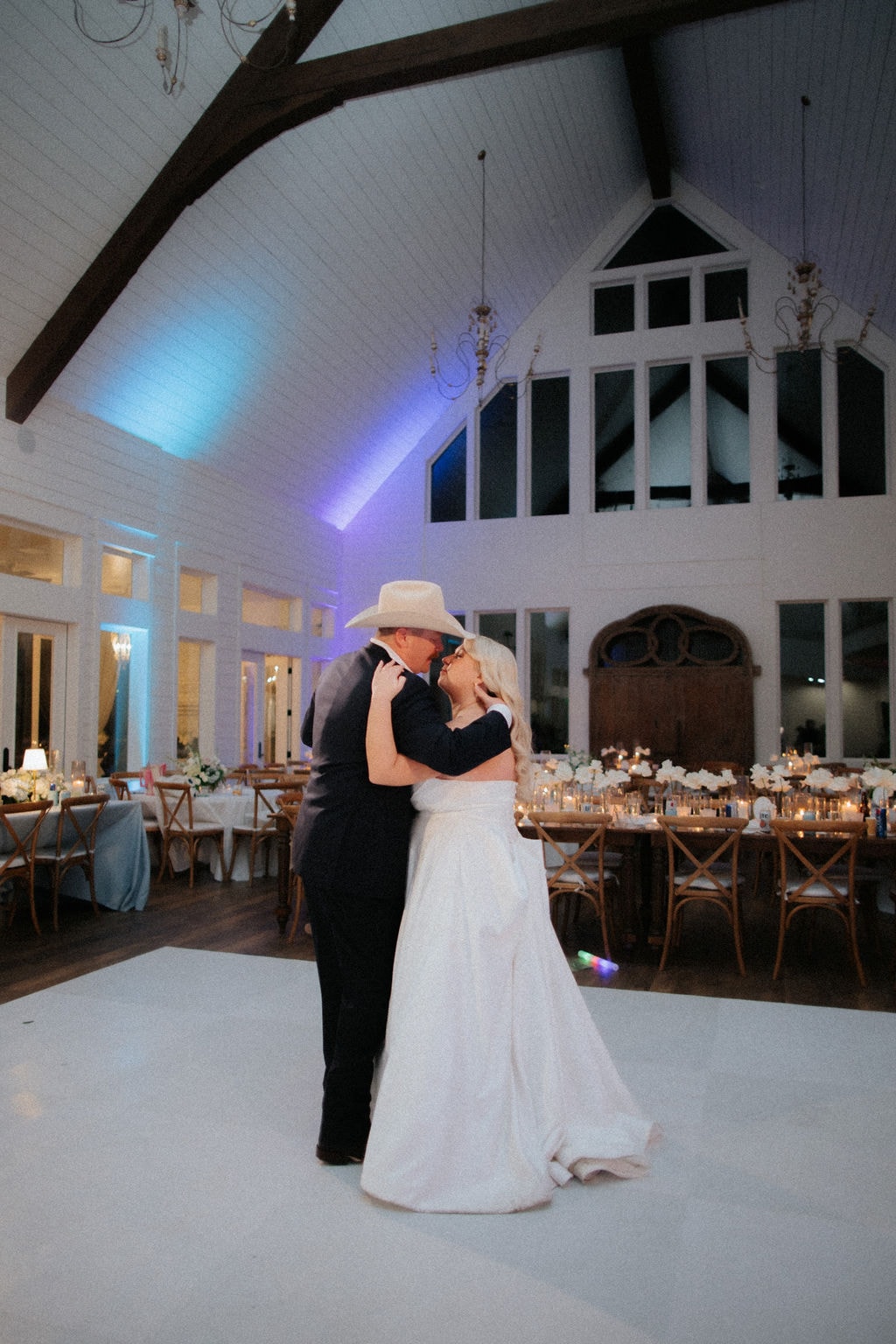 Kara and Anthony Marquardt sharing their first dance on the white Inkredible dance floor at The French Farmhouse Venue — Anthony in his cream cowboy hat, blue uplighting glowing across the soaring cathedral ceiling