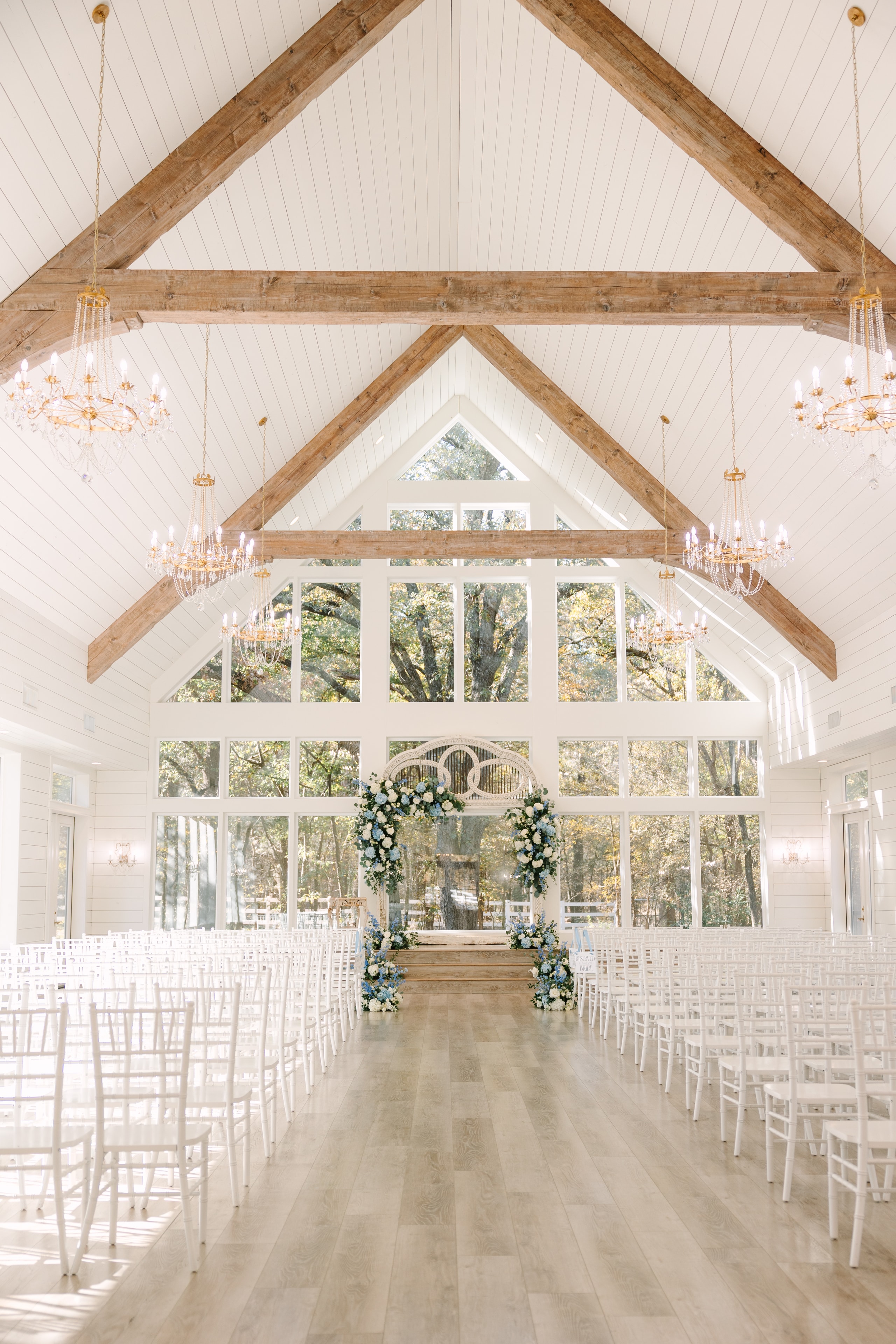 The French Farmhouse chapel interior set for Katie and Mason's ceremony — soaring wood beam ceilings, crystal chandeliers, white chiavari chairs, and a blue and white floral arch at the altar with floor-to-ceiling windows overlooking the fall trees