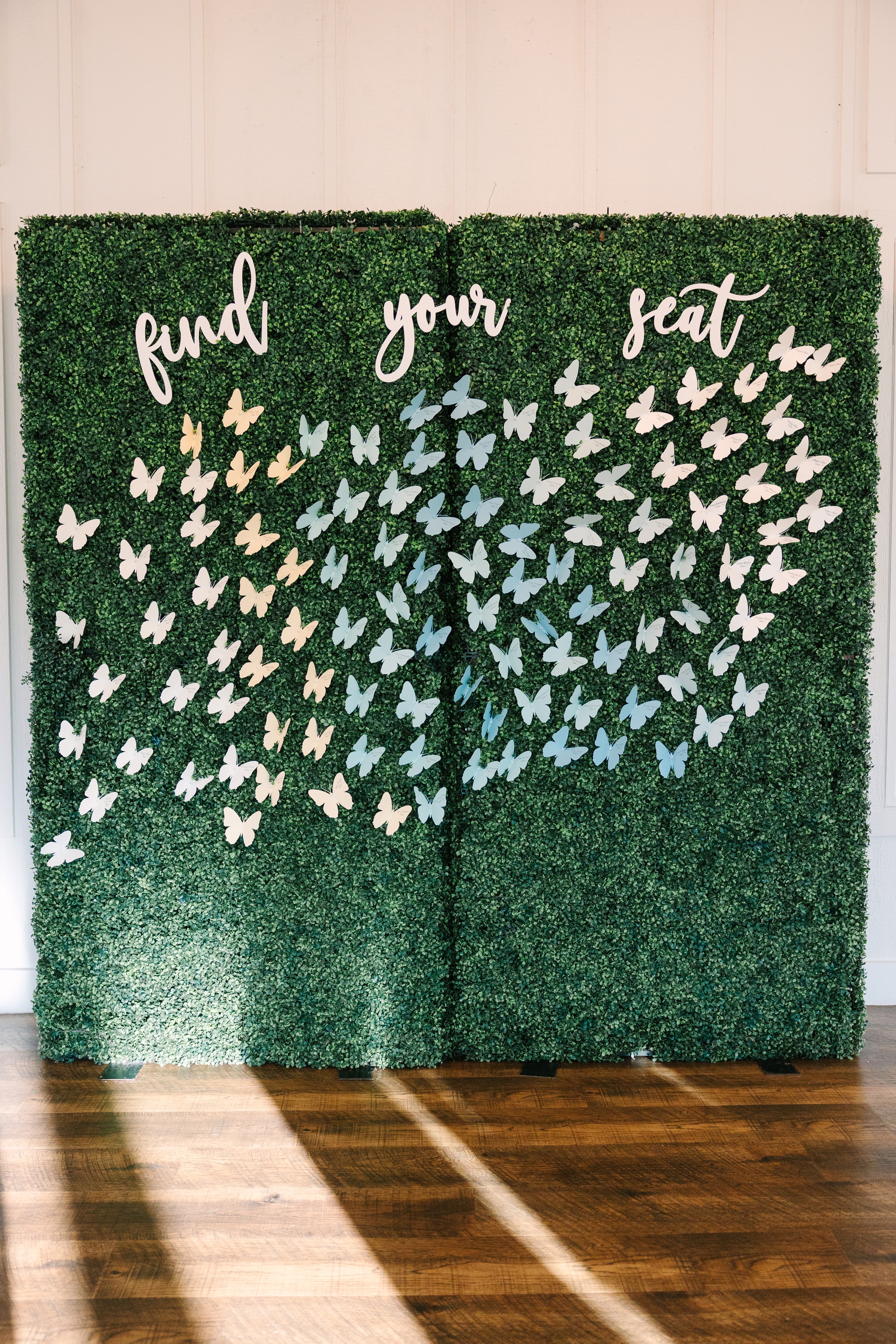 Katie and Mason's butterfly seating chart — a lush green hedge wall covered in white and blue paper butterflies with 'find your seat' in script lettering