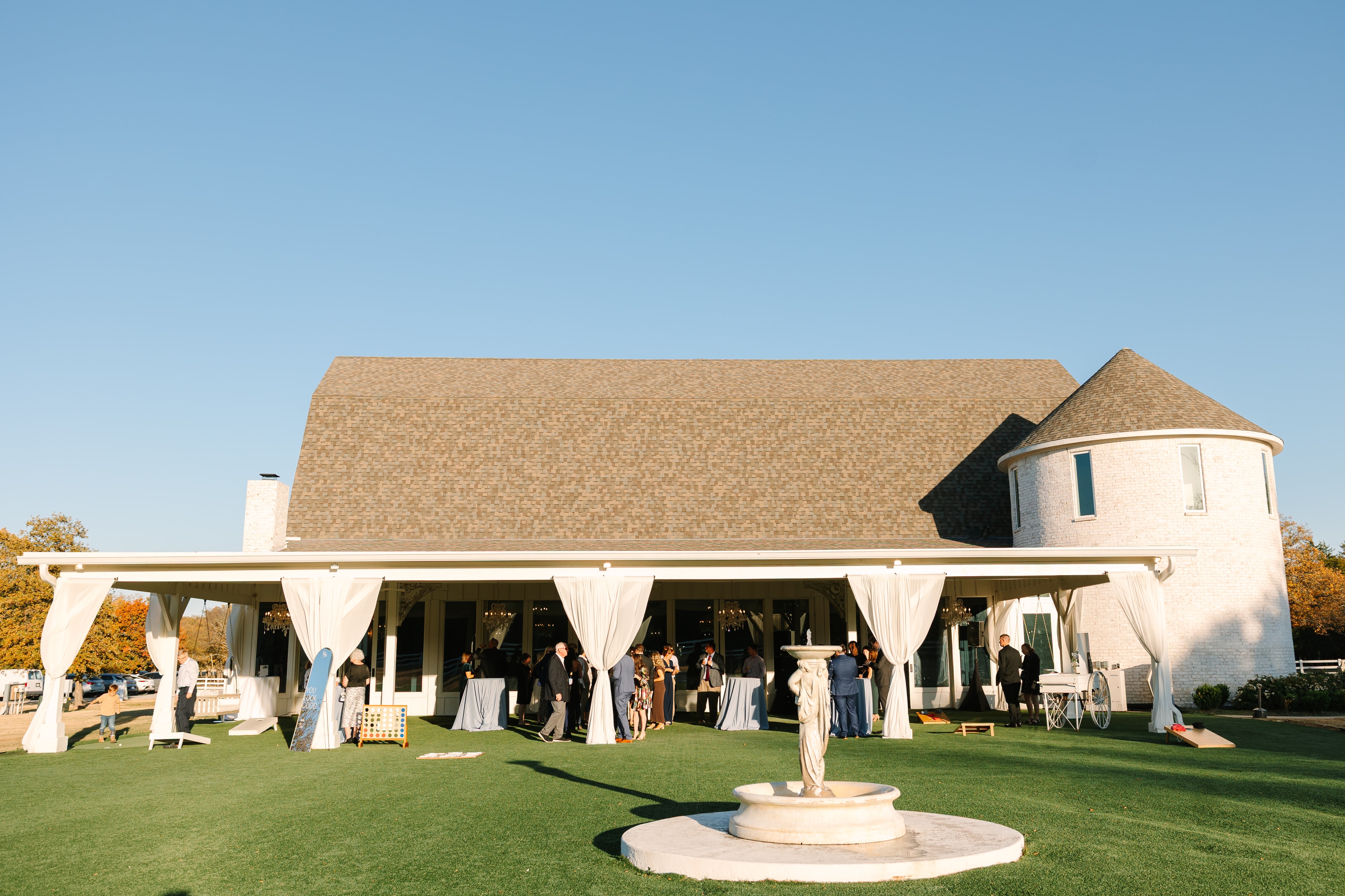 Guests mingling on the manicured lawn during cocktail hour at The French Farmhouse Venue — white draped pavilion, lawn games, and the iconic white farmhouse exterior with fall foliage in the background