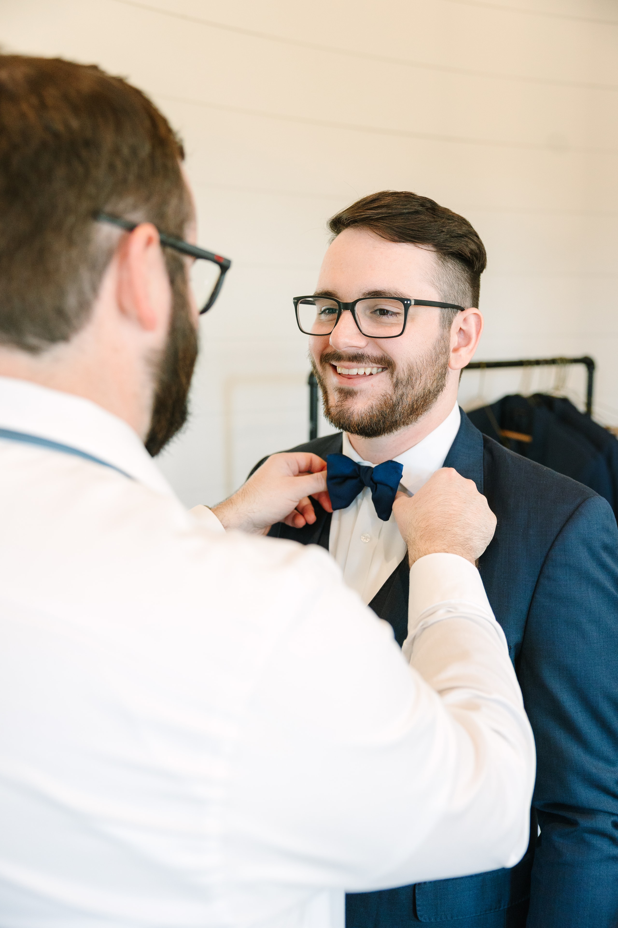 Mason smiling as a groomsman adjusts his bow tie in the groom's room at The French Farmhouse Venue