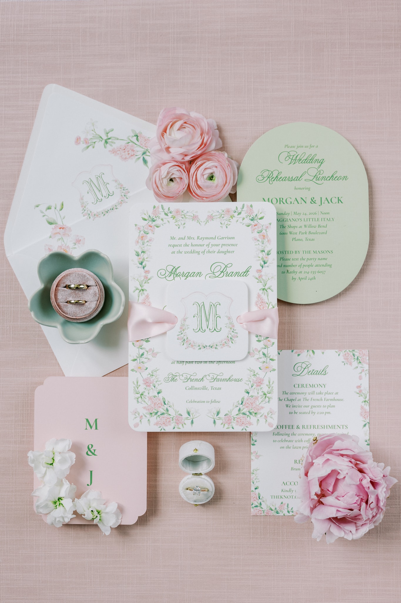 Morgan and Jack's wedding invitation suite flatlay — floral-bordered invitation with pink satin ribbon, sage green rehearsal luncheon card, rings in a mint ceramic dish, and pink ranunculus and peony blooms