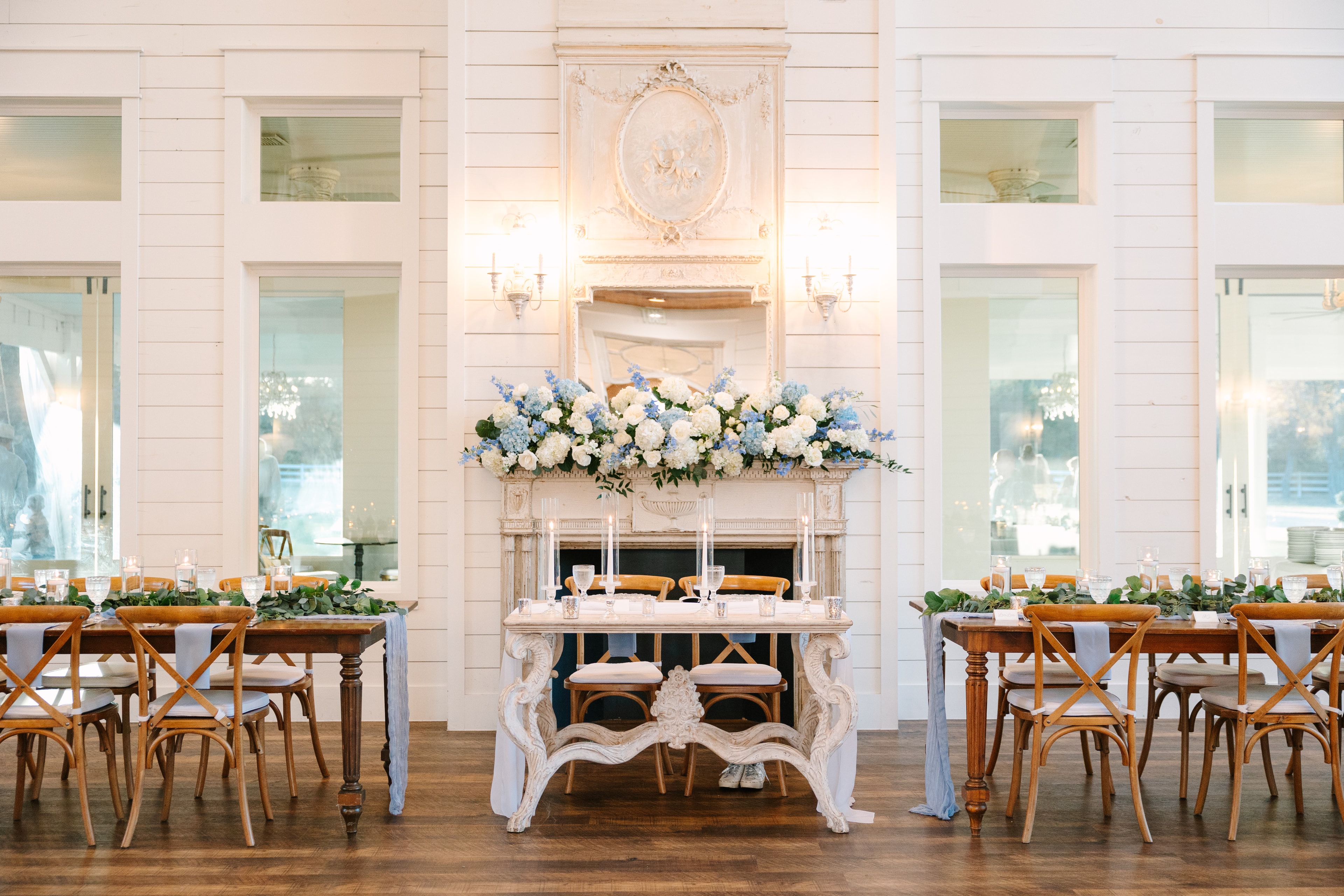 Sweetheart table in front of the ornate fireplace mantel adorned with blue hydrangeas and white roses at The French Farmhouse Venue reception hall