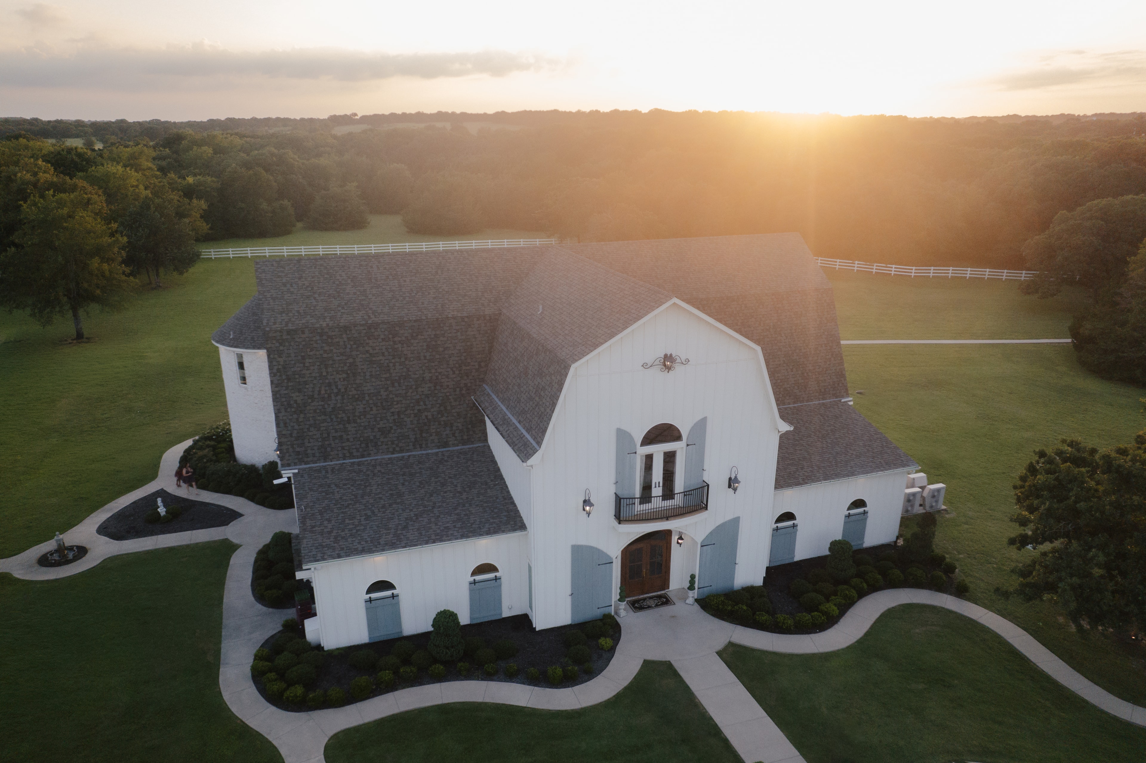 Aerial sunset view of The French Farmhouse Venue — white barn estate surrounded by lush green grounds and rolling Texas countryside at golden hour