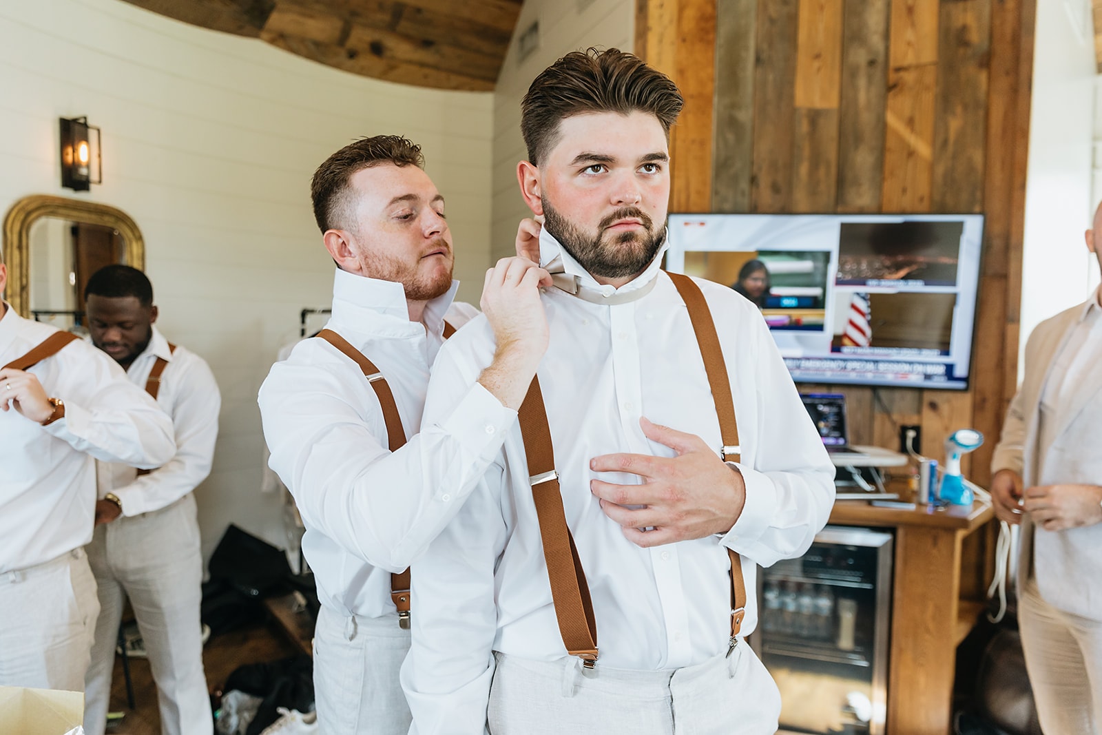 Groomsman helping the groom adjust his bowtie while wearing white shirts and brown suspenders