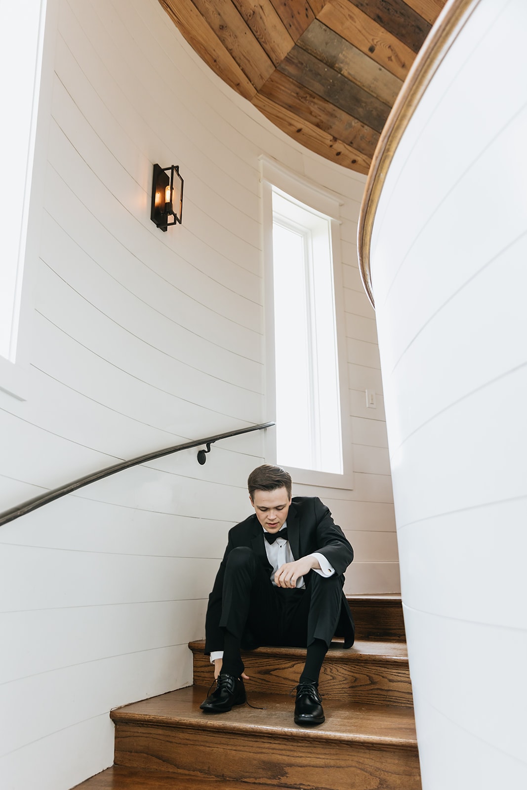 Groom in full tuxedo sitting on the spiral staircase tying his shoes